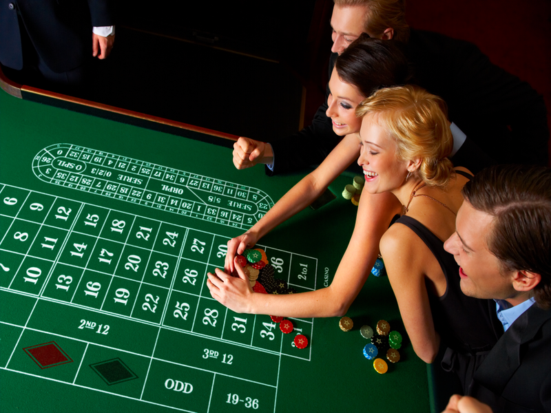 Zwei Damen und zwei Herren lehnen sich lachend über einen Roulettetisch. Eine der Dame schiebt einen Haufen Roulette-Chips zu sich heran.