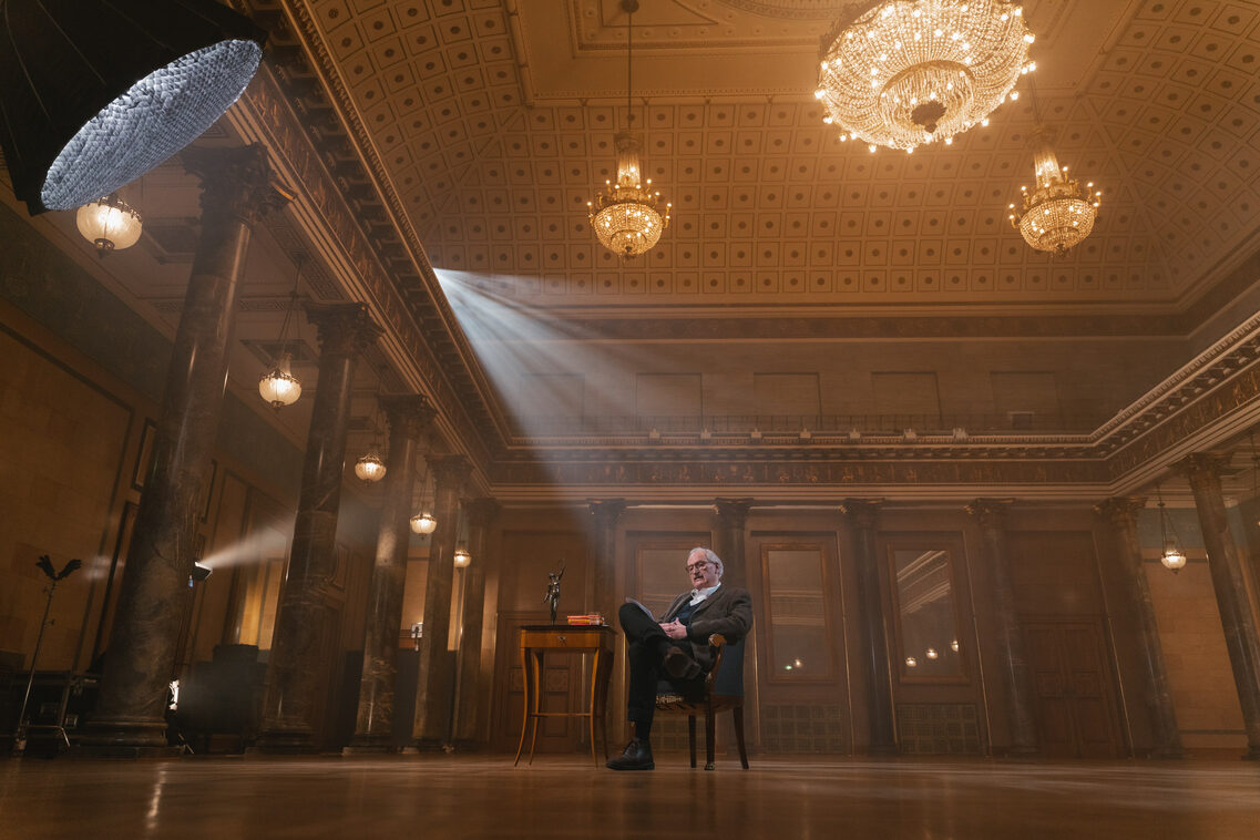 A man sits on the chair in the middle of the room in the Kurhaus. On the left is a side table with a flower in a vase.