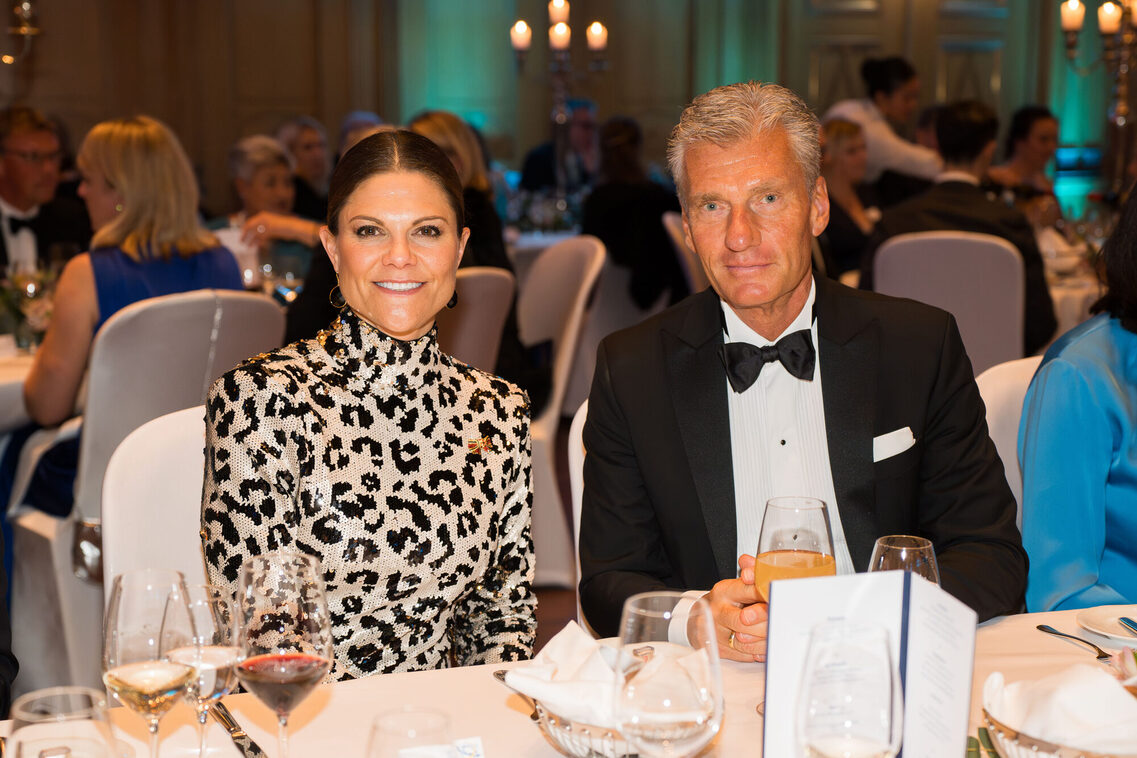 Eine Frau und ein Mann sitzen am Tisch, gucken in die Kamera und lächeln