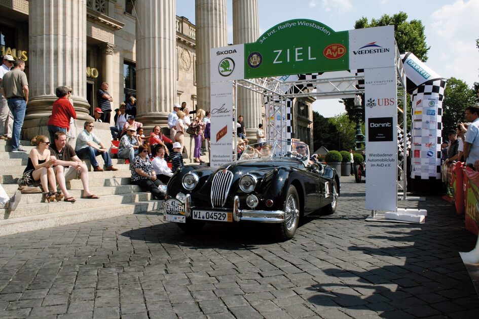 Vintage car drives through the finish arch on the Kurhaus forecourt. The audience sits and stands on both sides.