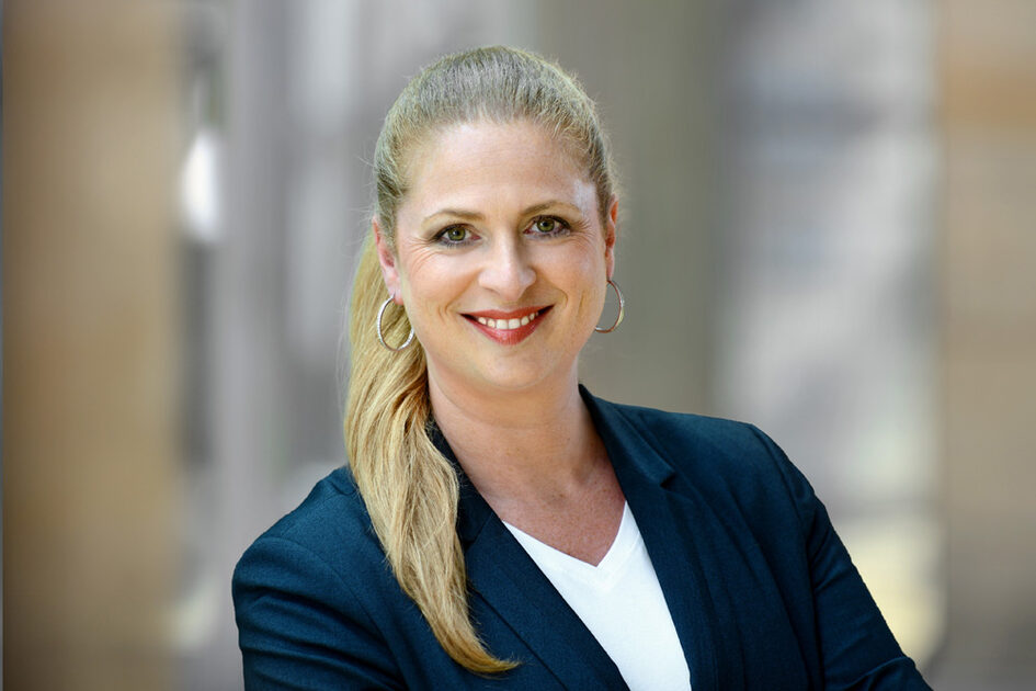 Portrait photo Tanja Volk smiling with her hair tied back in a ponytail and creoles in a blazer
