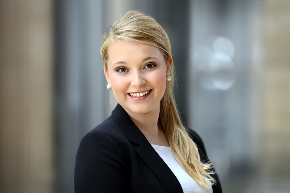 Portrait photo of Anna Apitz smiling with her hair tied back in a ponytail and wearing pearl earrings in a blazer.