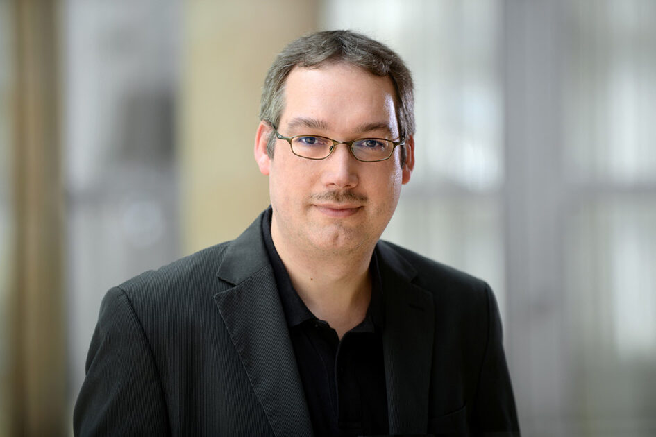 Porträtfoto Dirk Wolf mit Kurzhaarfrisur und schmalem Oberlippenbart. Er trägt eine schlichte Brille und Sakko mit Hemd.