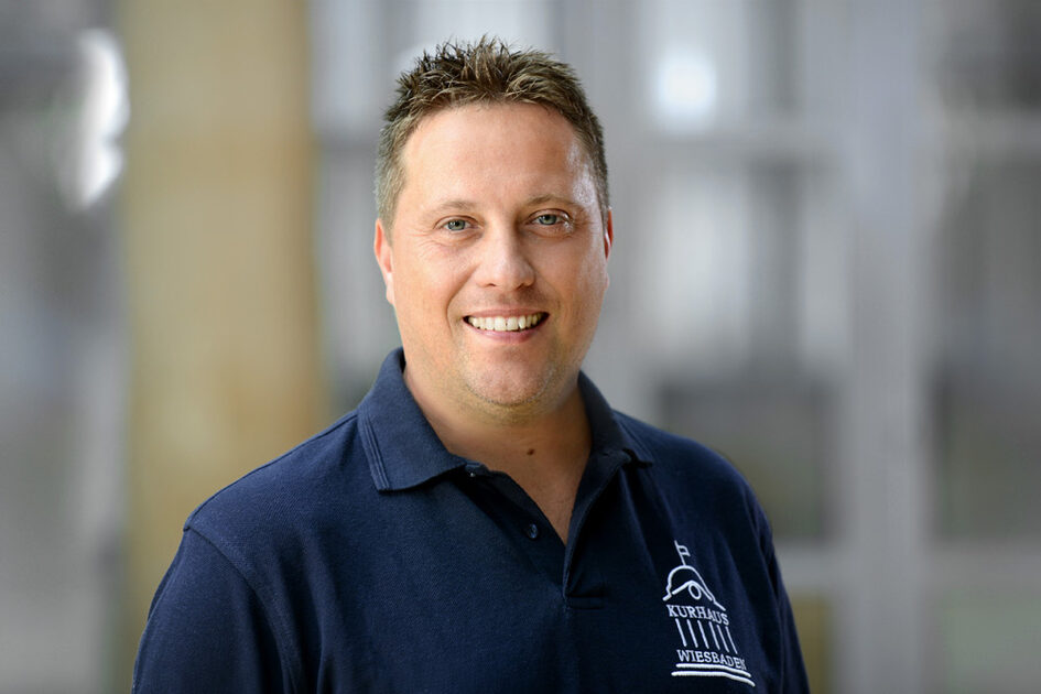 Porträtfoto Michael Hirschochs lächelnd mit Kurzhaarfrisur. Er trägt ein Poloshirt mit dem Kurhaus-Logo auf der linken Brust.