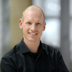 Porträtfoto Hagen Flick lächelnd mit Halbglatze. Er trägt ein Hemd.