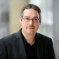 Porträtfoto Dirk Wolf mit Kurzhaarfrisur und schmalem Oberlippenbart. Er trägt eine schlichte Brille und Sakko mit Hemd.