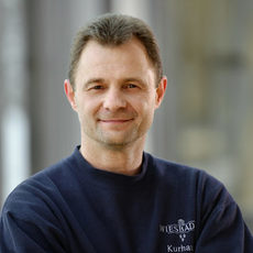 Porträtfoto Ralf Thiem lächelnd mit Kurzhaarfrisur. Er trägt einen Pullover mit Kurhaus-Aufschrift auf der linken Brust.