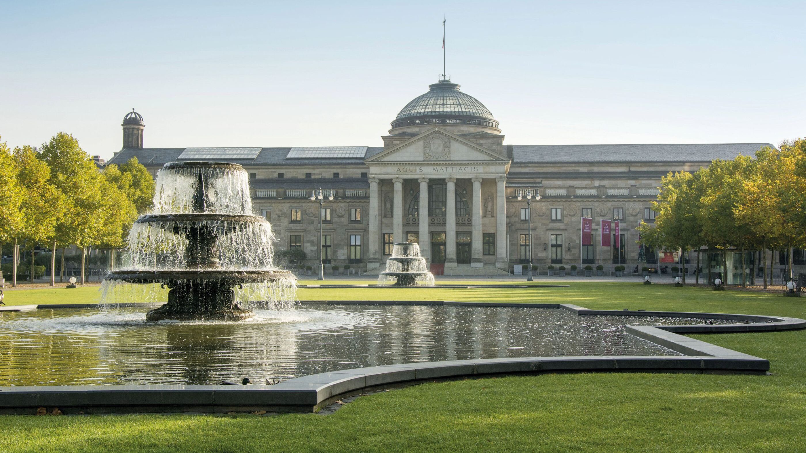 Kurhaus Wiesbaden mit von Bäumen gesäumtem Bowling Green mit zwei Brunnen.